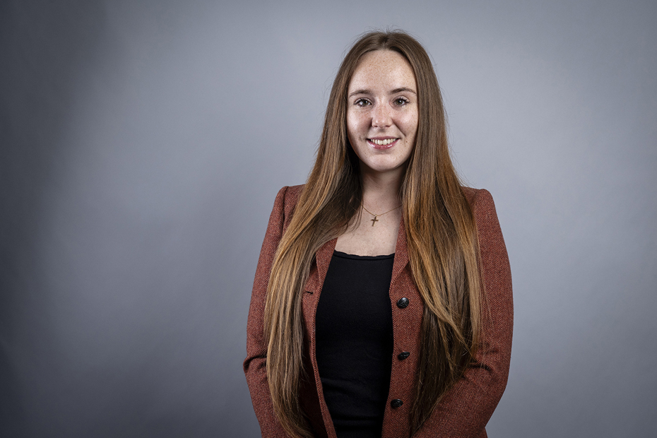 Professional headshot of a Solicitor in a black top and orange jacket, standing against a grey background.